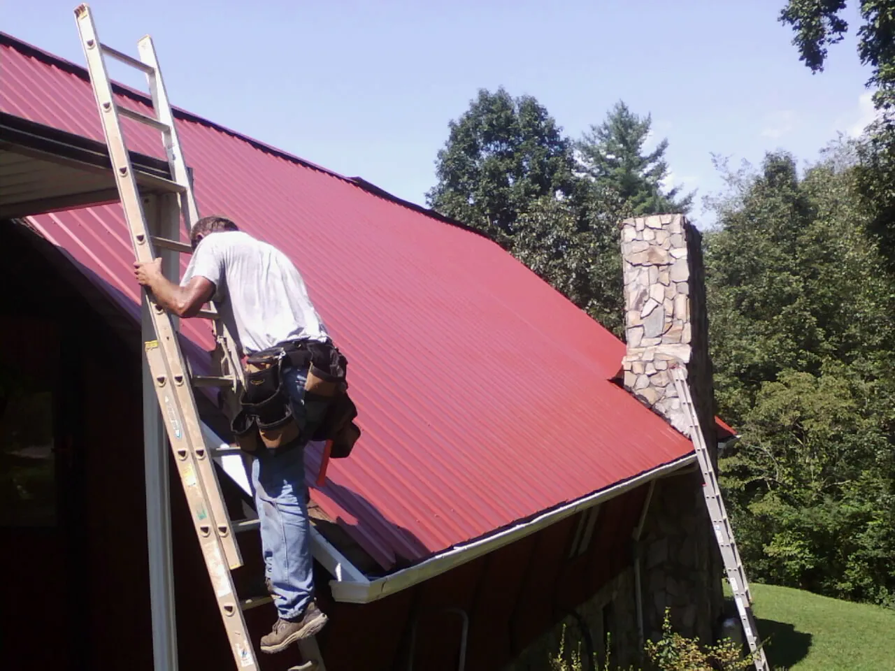 Roofer on a ladder beside a red metal roof and stone chimney — Soffit & Fascia Repair by Roofing Brothers in Bondsville